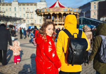 Happy young couple in love walking city street at bright sunny day, wearning in bright yellow and red down jackets. Pretty girl turn around and looking at camera. lifestyle portrait. Romantic, love and Valentine's Day concept.の写真素材