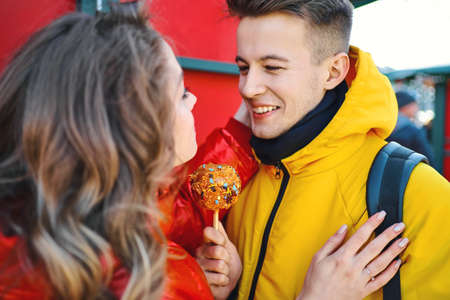 closeup portrait young smiling man in yellow down jacket holding sweets and looking at his girlfriend on red background. beautiful couple in love walking city sunny day and enjoying together Valentines day holiday.の写真素材