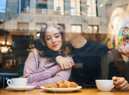 young Happy romantic couple sitting over the window in cafe, embracing, kissing and drinking coffee. boyfriend tenderly hugging and kissing girlfriend in cozy cafe. View through cafe window. Romantic, love and Valentine's Day concept.の写真素材