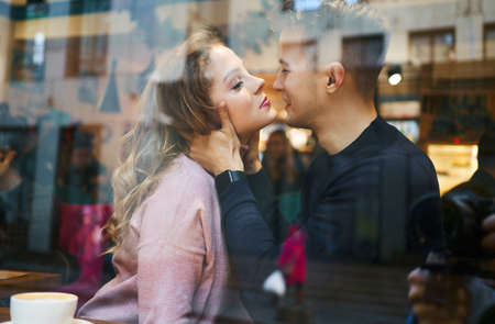 young romantic couple sitting over the window in cafe, embracing and kissing. Cute boyfriend and girlfriend having a date, enjoying together Valentines day holiday. View through cafe window.の写真素材