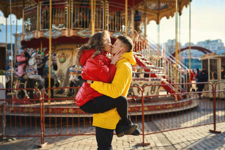 young romantic couple wearing in bright yellow and red down jackets walking city at sunny day, kissing, hugging. Handsome man holding young attractive woman on french carousel background. Romantic, love and Valentine's Day concept.の写真素材
