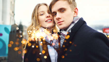 portrait of elegant romantic couple smiling to the camera, with sparkle in hands on festive city decorations background. Romantic, love and Valentine's Day concept.の写真素材