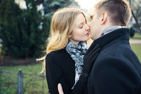 portrait of beautiful elegant woman in black coat kissing man with closed eyes on green park background, happy moments together. Romantic, love and Valentine's Day concept.の写真素材