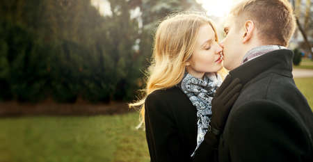 portrait of beautiful elegant woman in black coat kissing man with closed eyes on green park background, happy moments together. Romantic, love and Valentine's Day concept.の写真素材
