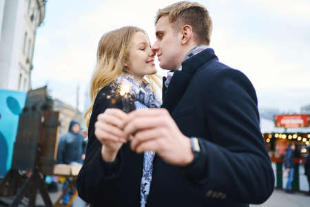 elegant blonde couple with sparkle in hands hugging and kissing on festive city decorations background. Romantic, love and Valentine's Day concept.の写真素材