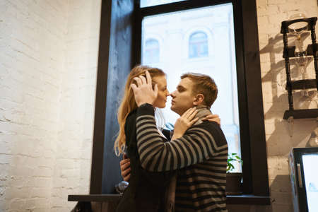 romantic couple in love sitting over the window in cafe on a date. portrait of attractive blonde woman and man embracing and touching each other. Concept of first kiss, tenderness and love.の写真素材