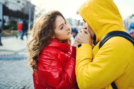 close-up portrait young tenderly romantic couple looking at each other, wearning in bright yellow and red down jackets. young man tenderly warms up hands his girlfriend. having a date. urban romantic portrait. Romantic, love and Valentine's Day concept.の写真素材