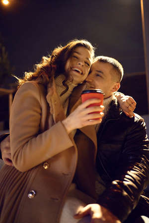 romantic couple in warm clothes embracing, laughing, looking happy, holding cups with hot drinks. having a date. urban romantic portrait. Romantic, love and Valentines Day concept.の写真素材
