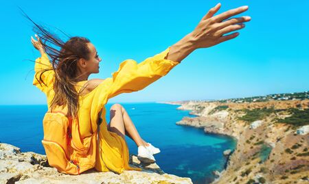 Close up portrait of taned traveler woman sitting on top rock beachwith raised hands in front of amazing seascape, bright yellow dress and hair blowing in the wind. Freedom, travel and vacation concept.の写真素材