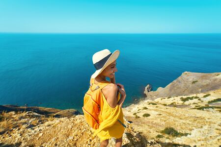 Bright portrait of happy smiling woman in bright yellow dress and hat with backpack standing on cliff edge against amazing seascape, admiring beautiful scenery of natural environment. Traveler enjoying recreation time and travel.の写真素材