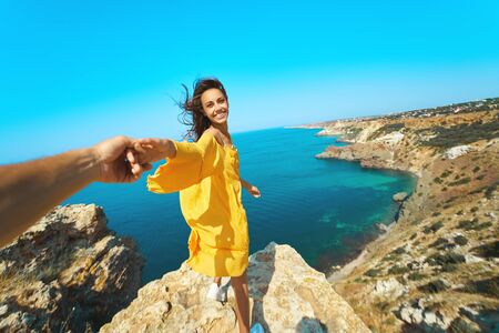 Portrait of happy carefree woman holding hand of her boyfriend, standing on cliff in front of amazing seascape, bright yellow dress and hair blowing in the wind. Follow me, travel, vacation concept.の写真素材
