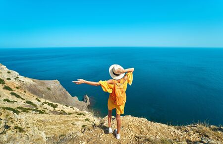 Back view of woman traveler in bright yellow dress and hat with backpack standing the top of mountain with raised hands against amazing seascape, admiring beautiful scenery of natural environmentの写真素材