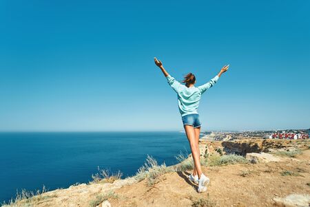 back view of carefree young woman standing with raised hands on cliff edge with beautiful sea view and enjoying wonderful nature. Caucasian female tourist having a great time on her vacation. Full length portraitの写真素材
