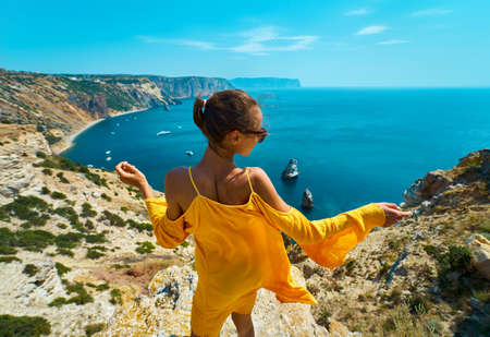 back view of taned traveler woman standing with arms spread open on cliff edge in front of amazing seascape,posing in bright yellow dress blowing in the wind. Freedom, travel and vacation concept.の写真素材