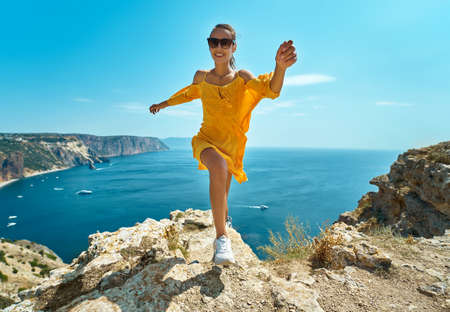 Happy tanned woman running on on cliff against amazing seascape, seashore. Smiling girl in sunglasses and bright yellow dress blowing in wind. Active and travel lifestyle concept.の写真素材