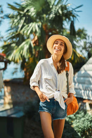 Carefree female tourist in straw hat, white shirt and orange backpack on vacation in Thailand, smiling and enjoying solo travel, sightseeing city street. Tourism and travel concept.の写真素材