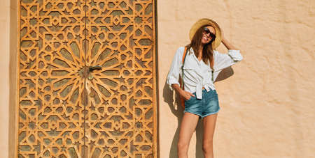 smiling tourist woman in straw hat, white shirt and sunglasses, standing against bright orange wall and authentic carved gate with traditional oriental ornament. enjoying summer vacation in Istanbulの写真素材