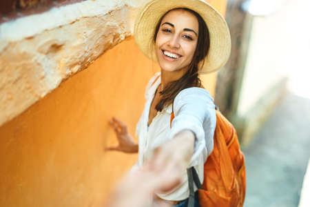 happy carefree tourist woman in straw hat, white shirt and orange backpack holding hand of her boyfriend, walking narrow street with bright orange walls. Summer vacation in Europe, Mediterranean. follow meの写真素材