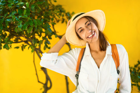 Close-up portrait happy woman tourist in straw hat, white shirt smiling and looking to camera, bright yellow wall on background. exploring old town streets. Vacation, tourism and travel concept.の写真素材