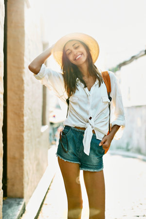 portrait carefree female tourist standing on ancient street with sunlight on bakcground. Attractive woman in straw hat, white shirt and sunglasses enjoying solo vacation in Europe. Tourism and travel concept.の写真素材