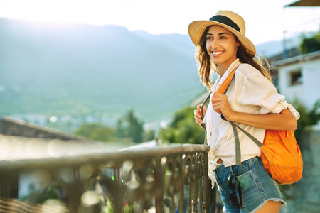 Portrait beautiful tourist woman standing on viewpoint with mountains and sunlight on bakcground. female in hat, white shirt, orange backpack enjoying summer adventure vacation in Europe.の写真素材