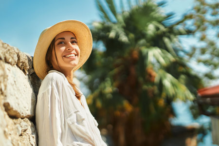 Portrait joyful woman tourist in straw hat, white shirt standing by old stone wall on vacation in Thailand, smiling and enjoying travel, sightseeing city street. Tourism and travel concept.の写真素材
