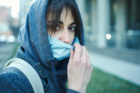 Coronavirus pandemic. Close-up portrait woman in hood wearing medical face mask, looking to camera on urban background. Prevention of bacterial infection, Corona virus, Covid 19.の写真素材