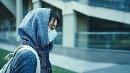 side view woman in hood with face mask for protection of coronavirus with modern building on background. Concept coronavirus pandemic in city, SARS, MERS, COVID-19, quarantine, isolation period.の写真素材