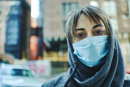 Coronavirus pandemic in city. Close-up portrait woman in hood with face mask looking to camera, standing in front of buildings on street. Prevention of bacterial infection, Corona virus, quarantine.の写真素材