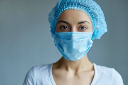 Portrait of a female doctor or nurse wearing medical cap and face mask looking at the camera on grey background. Ready to receive patients in hospital. Health care, medical concept.の写真素材