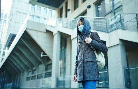 woman in hood with backpack standing against modern building in face mask for protection of coronavirus, looking to camera. concept coronavirus pandemic in city, SARS, MERS, COVID 19, quarantine.の写真素材