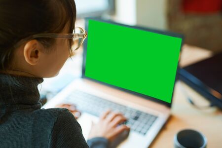 Back view woman freelancer in eyeglasses sitting at desk and typing on laptop with a green screen on monitor. Concept remote work, freelance, using laptop computer or net-book.の写真素材
