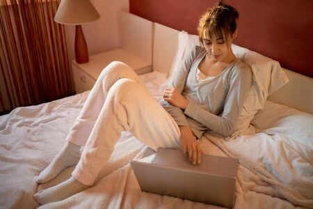 Young woman sitting on bed at home and watching laptop computer, browsing on Internet, reading news, searching information in social networks. Stay at home and quarantine concept.の写真素材