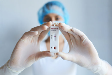 Doctor in protective face mask and gloves holding lab card kit test for viral disease COVID-19,...