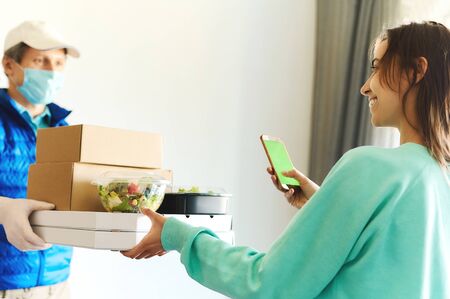 woman receiving packages from delivery man. Courier in uniform face mask gloves holding paper boxes for take out food and bags. Delivery food service at home, shopping order online from smartphoneの写真素材