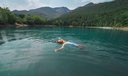 Traveler woman swimming on back in beautiful mountain lake with clear blue water against mountains. beautiful scenery, people in nature and travel conceptの写真素材