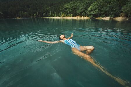 traveler carefree woman relaxing and floating in calm clear water, swimming on back in blue lake. Outdoor adventure, people in nature and calmness conceptの写真素材