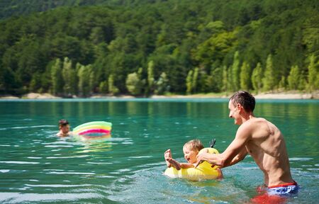 Athletic happy man having fun with kids in mountain lake with clear blue water. Family swimming, hot summer bathing, enjoing vacation outdoors. Family outdoor adventure, beautiful sceneryの写真素材