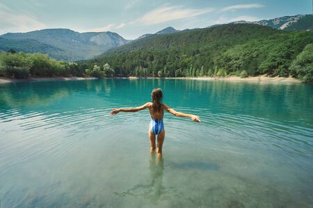 Back view freedom woman in swimsuit standing with open arms in mountain lake and mountains in background. inspiring landscape and beautiful scenery of nature.の写真素材