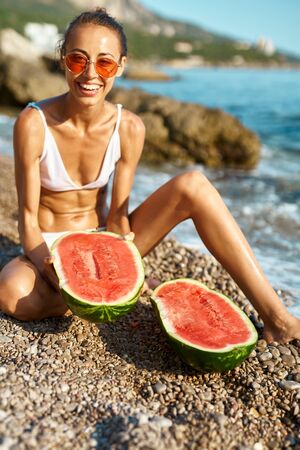 suntanned laughing woman in white bikini and red glasses sitts on pebble beach with two halves of huge ripe watermelon. focus on watermelonの写真素材