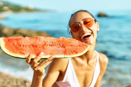joyful smiling girl in bikini holding huge slice of watermelon sitting on sea beach at seaside at summer vacation travel. focus on watermelonの写真素材