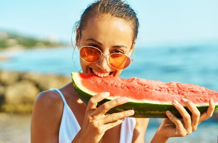 Close-up portrait summer woman in red glasses funny eating watermelon, bites him with pleasure on sea beach at summer vacation, enjoying juicy fruit.の写真素材