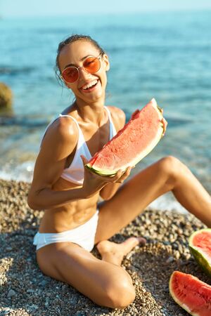 Tanned joyful girl in white bikini and sunglasses holding huge slice of watermelon sitting on sea beach at seaside at summer vacation travel, healthy food nutrition. focus on watermelonの写真素材