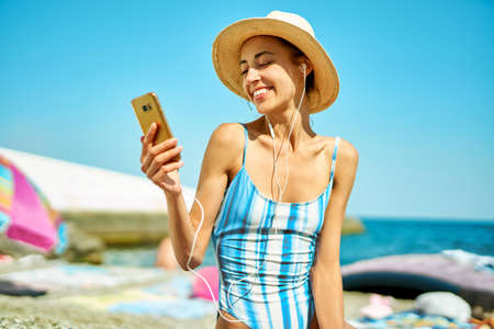 happy woman in straw hat and swimsuit at beach communicating on smartphone, online video cell phone call. Technology and communication during summer vacationの写真素材