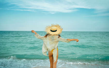 Beach summer stylish girl in happy freedom concept standing back with open arms by blue ocean with waves, enjoying sea breeze. Woman in big straw hat on windy beach, rear viewの写真素材