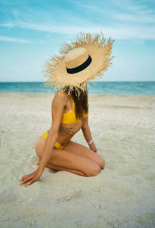 vertical shot sexy bikini tanned body woman wearing big straw hat sitting on sand beach by blue sea. Summer lifestyle portrait and summer beach concept, no faceの写真素材