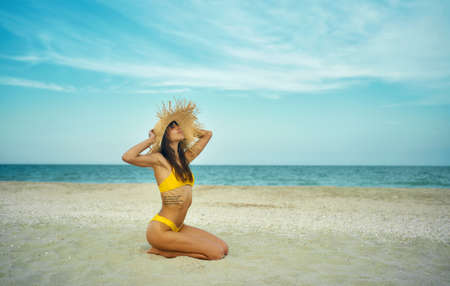 Beautiful tanned woman on tropical beach by ocean with copy space. girl wearing in yellow bikini and big straw hat sits on sand, breathing and enjoying sea breezeの写真素材