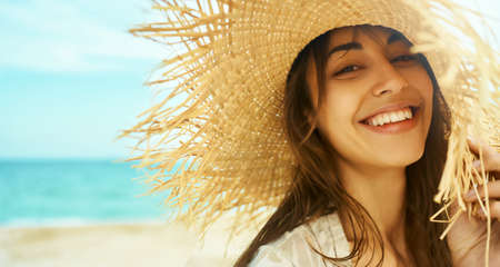 Close-up face smiling woman wearing straw hat at beach in sunshine sunrise on seashore. Happy tanned travel woman laughing and looking to camera. relaxing at beachの写真素材
