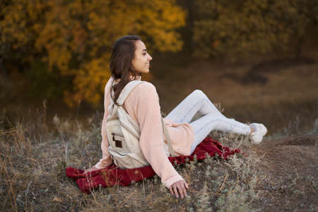 cozy portrait woman in warm sweater with backpack sits on blanket in autumn park with fall colorful background.の写真素材