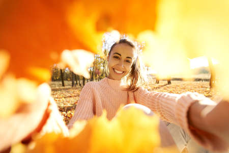 Delighted smiling girl makes selfie from frame witn fall leaves, enjoyng autumn sunny day at parkの写真素材